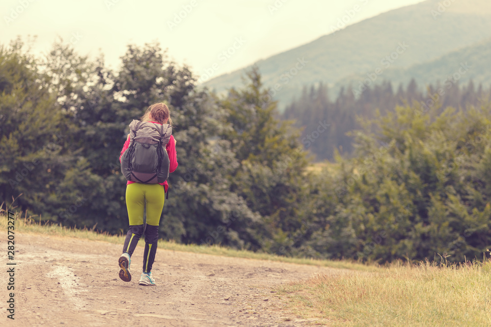 Woman with a backpack hiking in the forest on a bright sunny day. Rear view image of young woman carrying backpack walking through mountain trial on summer day. Woman with a backpack hiking in the forest on a bright sunny day. Rear view image of young woman carrying backpack walking through mountain trial on summer day.