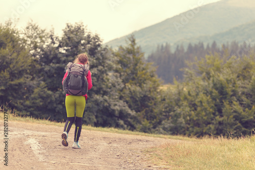 Woman with a backpack hiking in the forest on a bright sunny day. Rear view image of young woman carrying backpack walking through mountain trial on summer day.