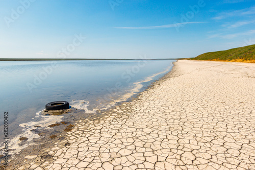 The shore of the blue lake with dirty water, dry cracked soil and discarded old used tire. Gruzskoe lake, Rostov-on-Don region, Russia