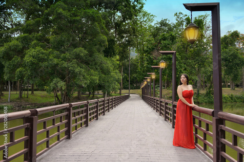 Woman in Red long dress standing at the bridge park in the evening with lamp ligths background
