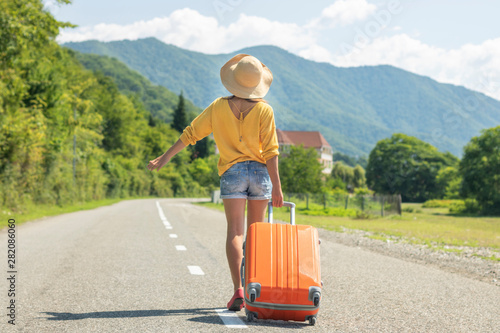Happy girl in the summer goes on vacation, on vacation with an orange suitcase on the background of nature and mountains. The concept of travel, weekend in a beautiful place, a house in the mountains