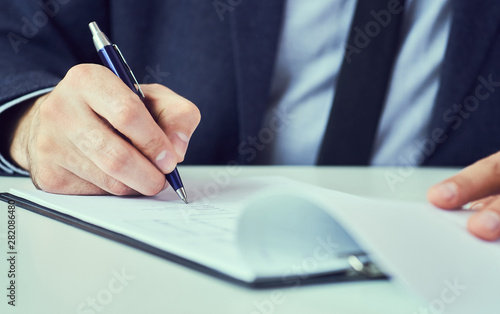 Hand of businessman in suit filling and signing with blue pen partnership agreement form clipped to pad closeup.