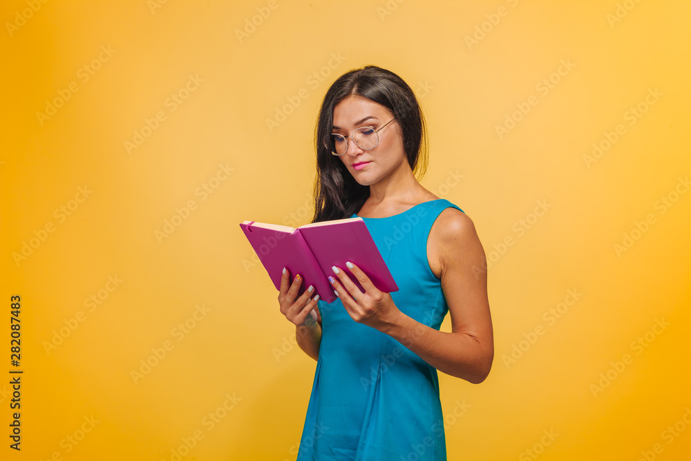 Girl with glasses on a yellow background in a blue dress reading a pink notebook Girl with glasses on a yellow background in a blue dress reading a pink notebook
