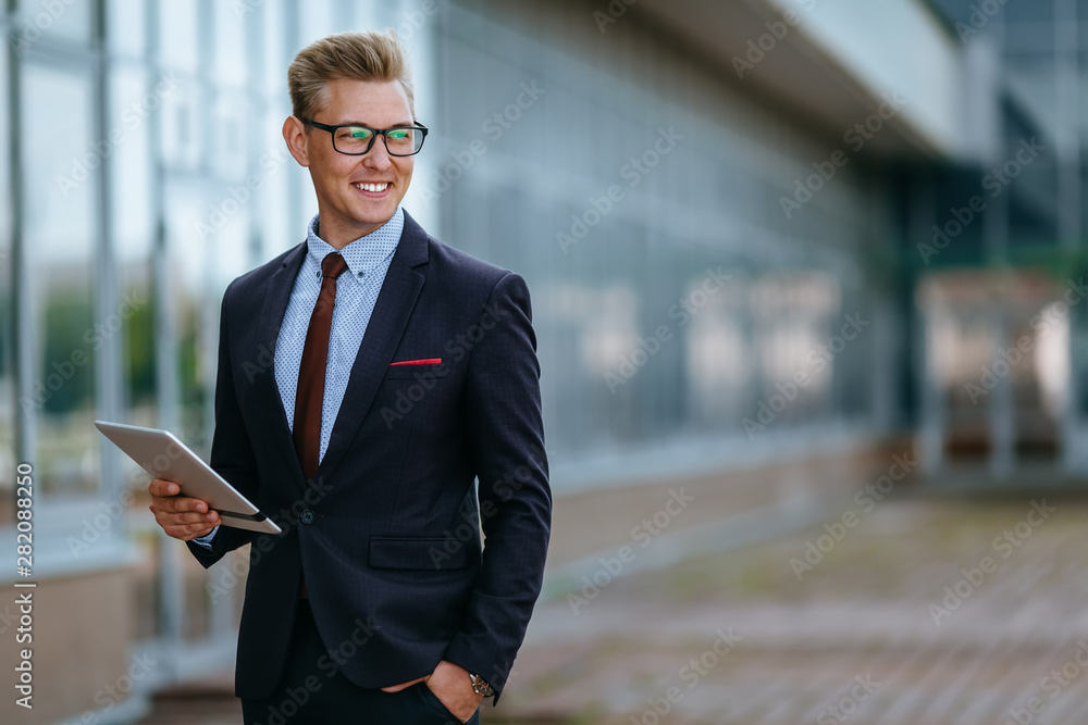 Elegant smiling young businessman using his PC tablet. Portrait of a Male executive manager in trendy clothes with tablet pc in front of office building. Modern technology concept Elegant smiling young businessman using his PC tablet. Portrait of a Male executive manager in trendy clothes with tablet pc in front of office building. Modern technology concept