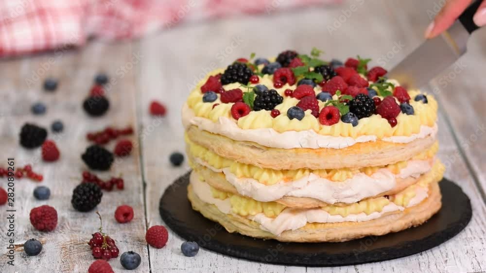Layered cake decorated with berries and mint on table. Woman cutting delicious puff pastry cake. Layered cake decorated with berries and mint on table. Woman cutting delicious puff pastry cake.