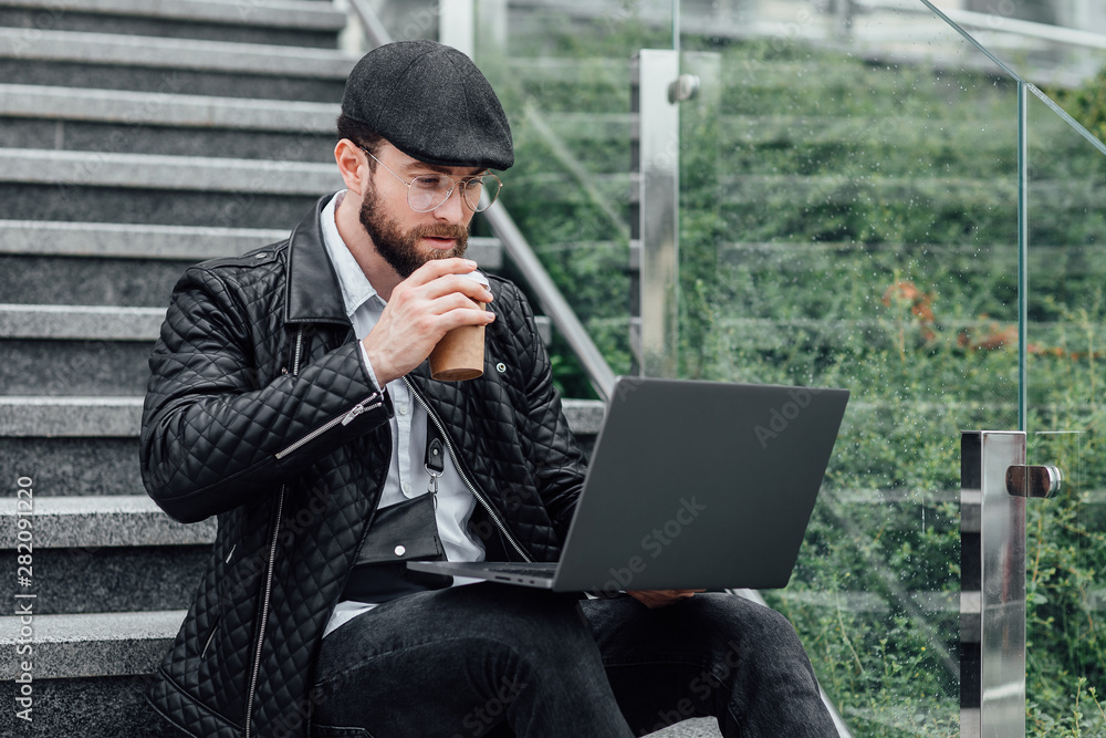 Handsome young manager man drinking coffee and working on laptop while sitting outdoors on the stairs. Business concept. Handsome young manager man drinking coffee and working on laptop while sitting outdoors on the stairs. Business concept.