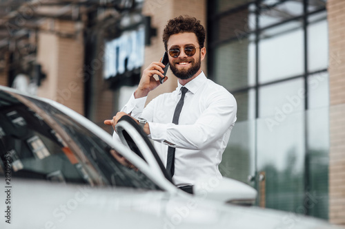 Handsome, smiling, bearded businessman in white shirt, speaking by phone and standing near his car outdoors on the streets of the city near the modern office center.