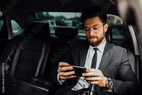 Handsome, bearded, smiling businessman in black suit surfing smart phone on backseat of the car. Texting to colleague.. © Тарас Нагирняк