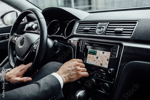 Closeup of man hands holding steering wheel and using build-in satellite navigator GPS in a new premium car. Searching new city on the map.