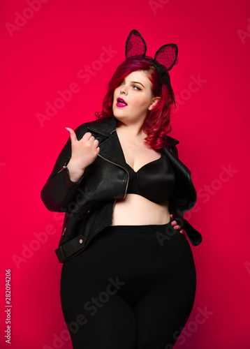 Cute sexy plus size brunette with black bunny ears in leather jacket and underwear posing on red background
