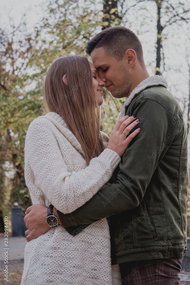 Couple in love stand on autumn fallen leaves in a park, enjoying a beautiful autumn day. Man hugs girl Couple in love stand on autumn fallen leaves in a park, enjoying a beautiful autumn day. Man hugs girl