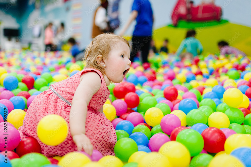 Portrait little cute baby girl princess infant 1-2 year standing and play with balloons, colorful balls in playground, ball pit, dry pool for birthday party. Celebration first year concept. Portrait little cute baby girl princess infant 1-2 year standing and play with balloons, colorful balls in playground, ball pit, dry pool for birthday party. Celebration first year concept.