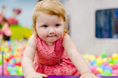 Portrait little cute baby girl princess infant 1-2 year standing and playing in the gaming room, with balloons, colorful balls in playground in birthday party. Celebration first year concept.
