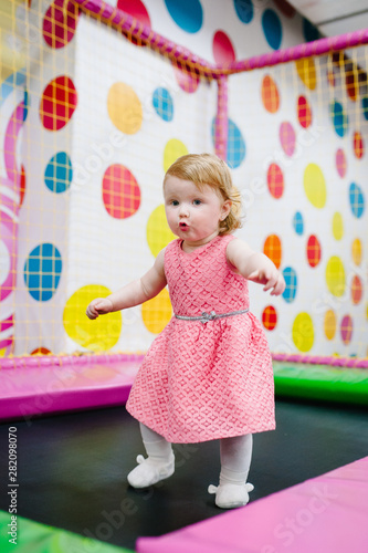 Portrait little cute baby child girl princess infant 1-2 year playing and jumping on a trampoline in children's playroom, indoor on birthday party. Celebration concept holiday, event, entertainment.