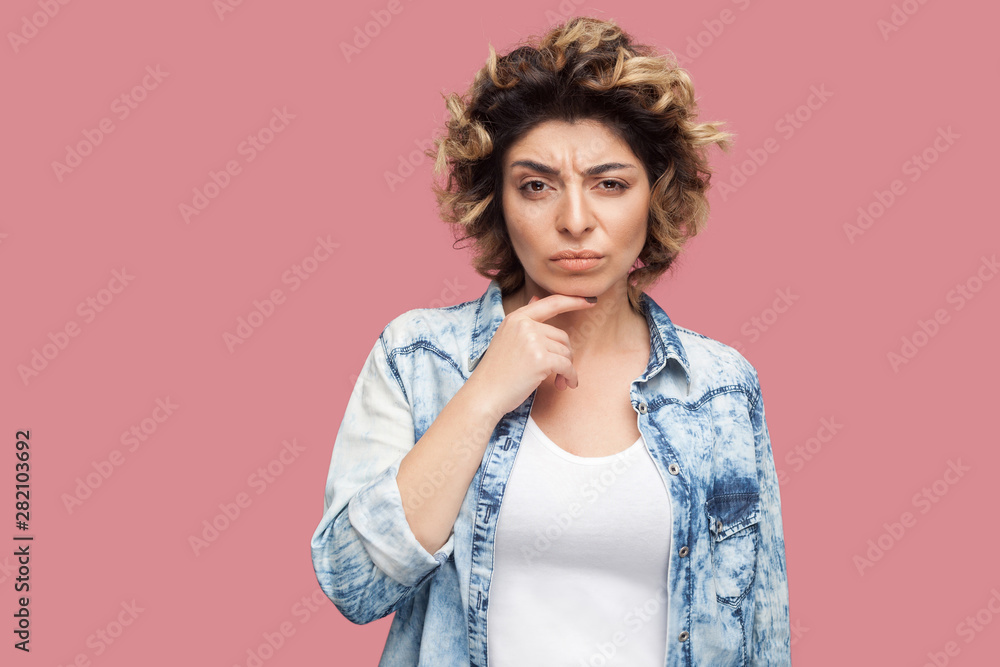 Portrait of thoughtful young woman with curly hairstyle in casual style standing, touching her chin and looking at camera with confused thinking face. indoor studio shot, isolated on pink background. Portrait of thoughtful young woman with curly hairstyle in casual style standing, touching her chin and looking at camera with confused thinking face. indoor studio shot, isolated on pink background.