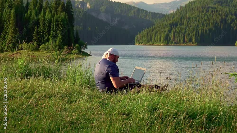 Mountain man working with laptop in the mountains, near mountain lake, with beautiful landscape on background Mountain man working with laptop in the mountains, near mountain lake, with beautiful landscape on background