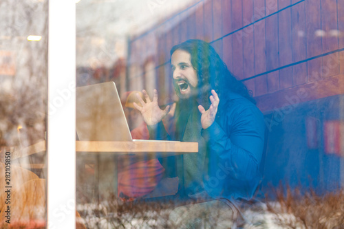Portrait through the window of angry aggressive young adult man freelancer in casual style sitting in cafe, trying to crash laptop screen, raised arms with clenching teeth. Outdoor, lifestyle concept