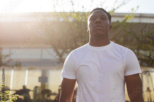 Back in lit muscular young man in white t-shirt training outdoors by gym in summer on blurred background. African American male fitness person working out by gym