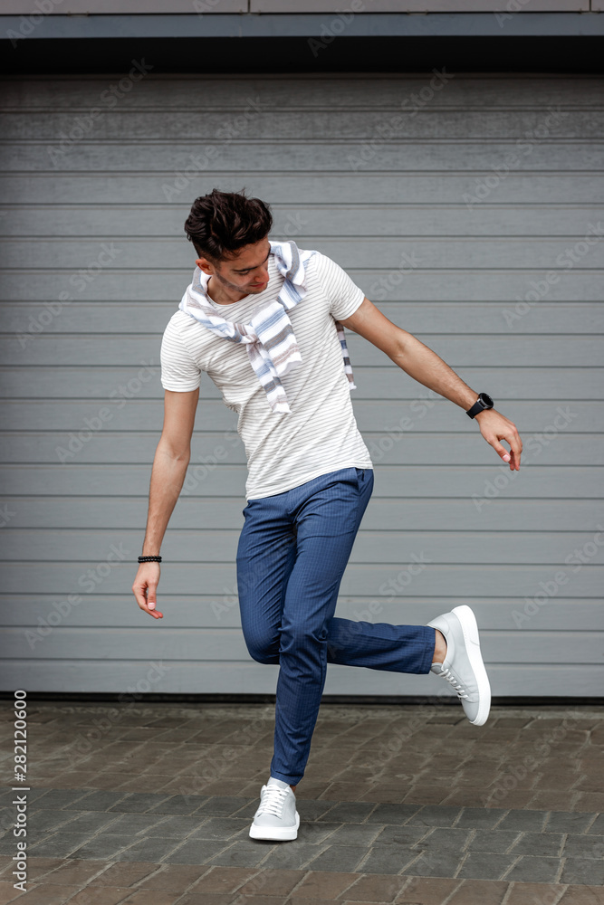 Stylish guy dressed in casual clothes trousers and t-shirt poses in the street on the background of a white fence