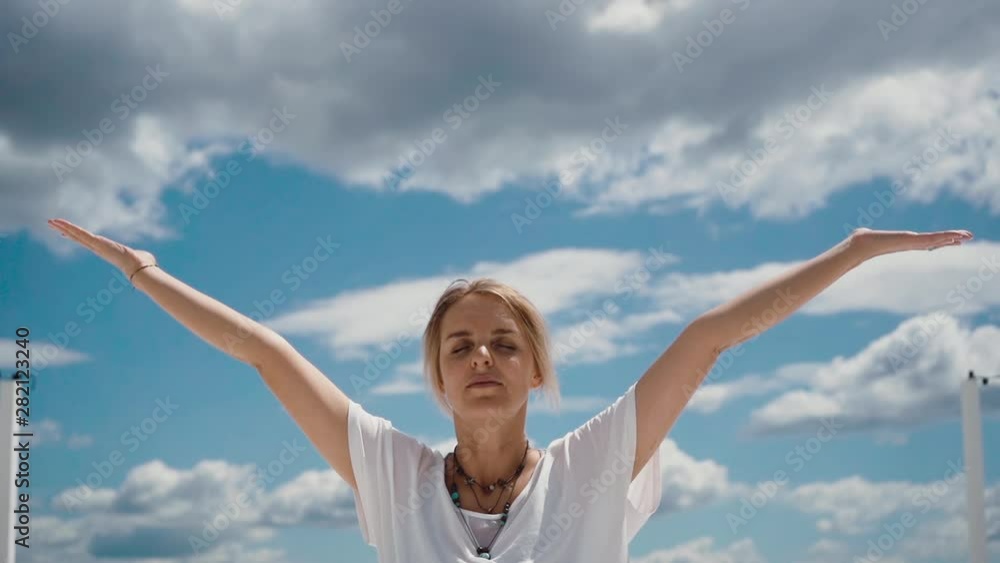Close-up portrait of beautiful young woman putting hands in namaste mudra posture. Girl meditates in background of white clouds and blue sky. Camera shoots from below in slow motion Close-up portrait of beautiful young woman putting hands in namaste mudra posture. Girl meditates in background of white clouds and blue sky. Camera shoots from below in slow motion