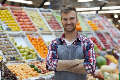 Waist up portrait of handsome young man working in supermarket and smiling at camera while posing by fruit stand, copy space photo