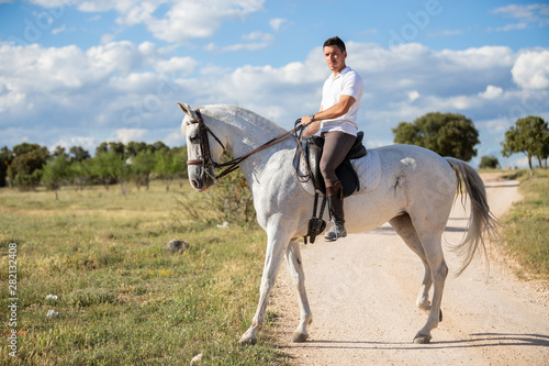 Young male in casual outfit riding white horse on grassy meadow a sunny day