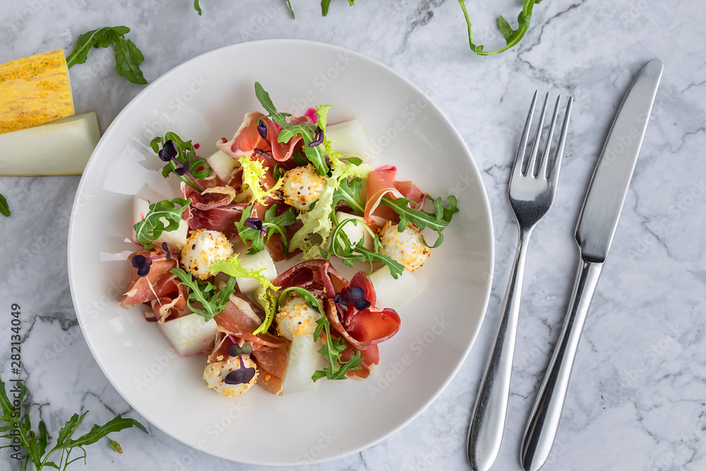 Salad with melon, proshuto, arugula and italian cheese in a white plate. Spoon, fork on the grey marble background Salad with melon, proshuto, arugula and italian cheese in a white plate. Spoon, fork on the grey marble background