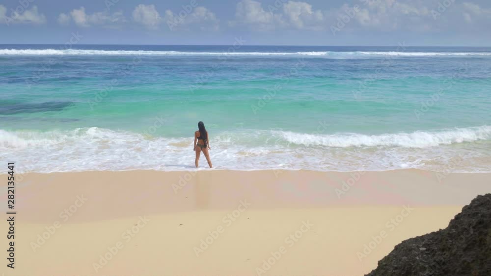 A panoramic shot of young asian woman with dark long hair is standing in water on tropical beach with bright sand in sunny day filmed in slow motion A panoramic shot of young asian woman with dark long hair is standing in water on tropical beach with bright sand in sunny day filmed in slow motion
