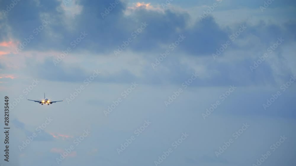 Small jet airplane with head lights is landing during cloudy weather with blue and yellow clouds in the sunset sky Small jet airplane with head lights is landing during cloudy weather with blue and yellow clouds in the sunset sky