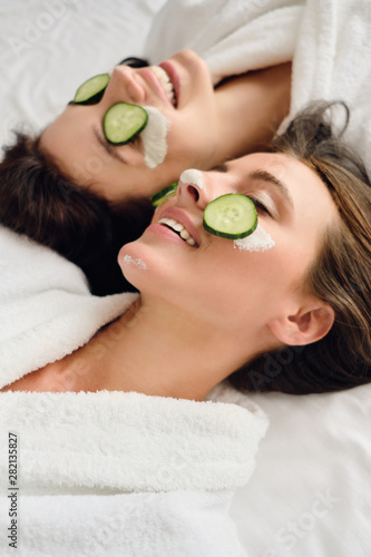 Two young attractive smiling women with dark hair in white bathrobes with cosmetic mask and slices of cucumber on faces happily lying in bed in modern hotel