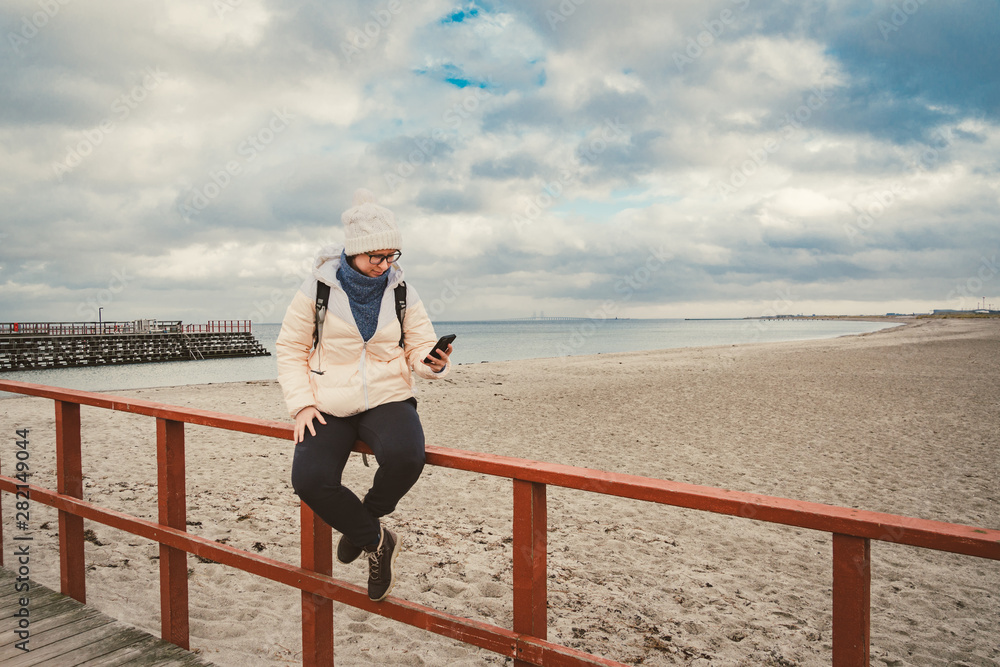 Caucasian woman in hat and jacket with a backpack in winter sits on a wooden pier on the beach near the North Sea. Denmark Copenhagen tourist uses a mobile phone on the shore. Scandinavia Travel Theme