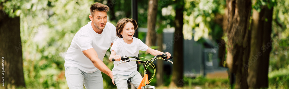 Obraz premium panoramic shot of father pushing bike while son riding bicycle