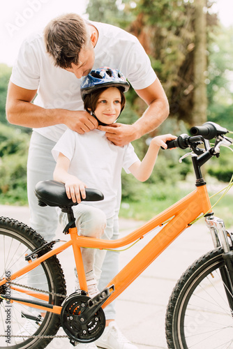 full length view of father putting helmet on son while boy standing near bicycle and looking away