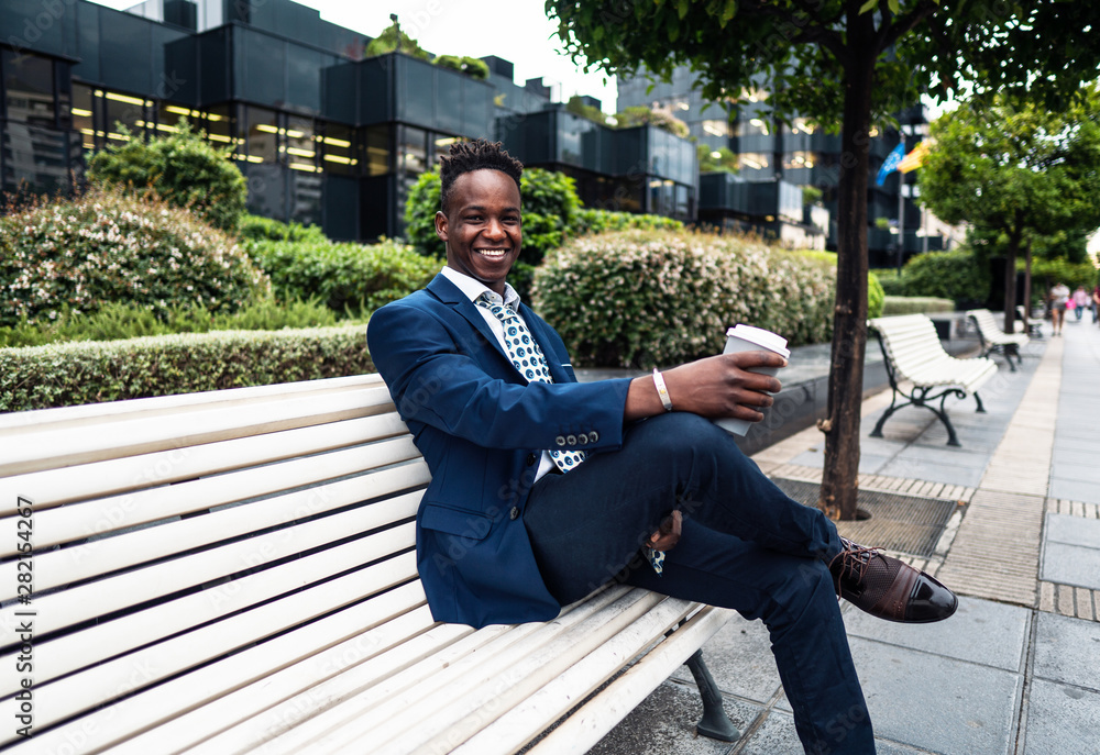 Obraz premium African American businessman wearing blue suit sits on the bench and drinks coffee near office. Business concept