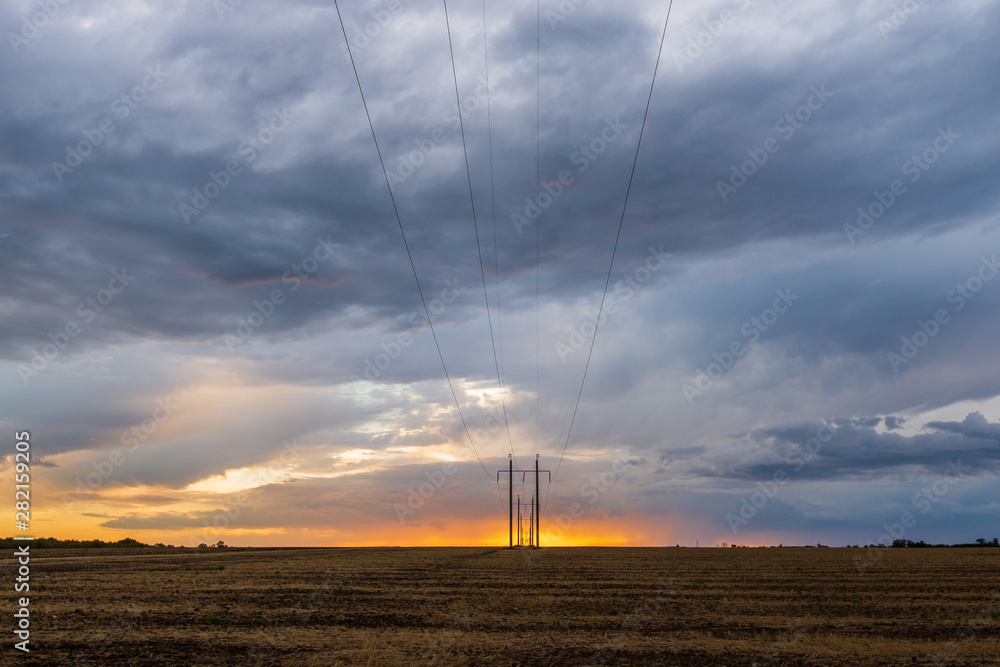 A row of high voltage electrical power lines cling to the horizon in an otherwise vast, wide open rural landscape on sunset with clouds