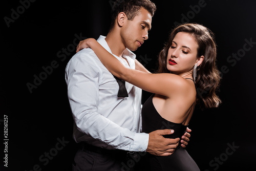 handsome mixed race man embracing elegant woman isolated on black