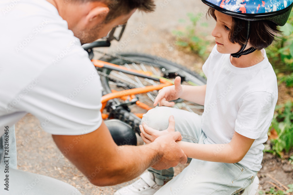 selective focus of father looking at knee of son because boy fell from bicycle