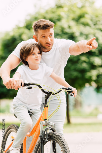 father and son looking forward while kid riding bicycle and dad pointing with finger