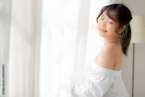 Portrait of beautiful young asian woman standing the window and smile while wake up with sunrise at morning, girl happy with freshness and cheerful, lifestyle and relax concept.