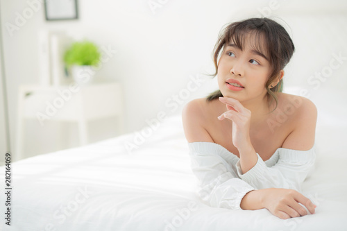 Beautiful portrait young asian woman lying and smile while wake up with sunrise at morning, beauty cute girl happy and cheerful resting on bed in the bedroom, lifestyle and relax concept.