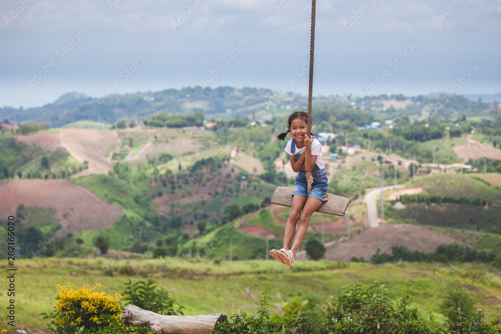 Happy asian child girl having fun to play on wooden swings in playground with beautiful nature