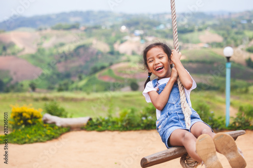 Happy asian child girl having fun to play on wooden swings in playground with beautiful nature photo