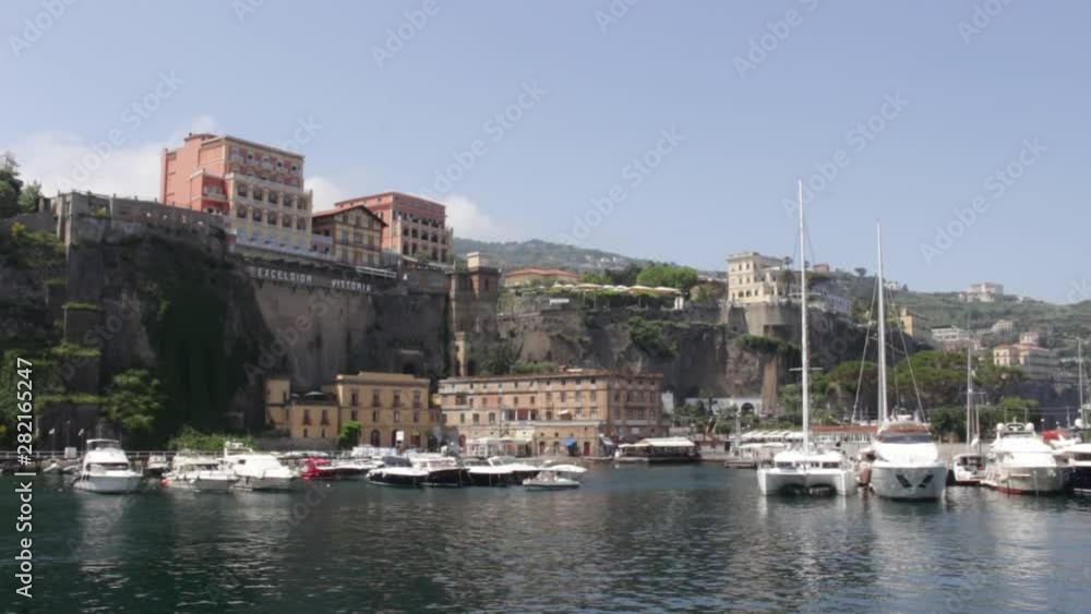 View of Boats Docked on Calm Water at Marina Next to Cliffside With Buildings and Mountains in Background in Sorrento, Italy View of Boats Docked on Calm Water at Marina Next to Cliffside With Buildings and Mountains in Background in Sorrento, Italy
