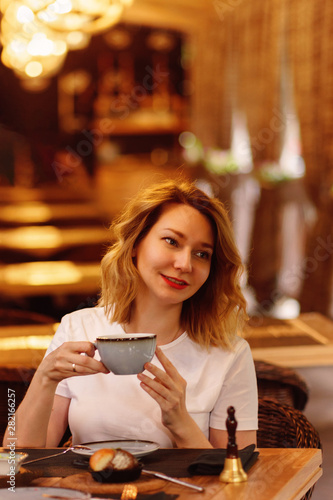 Pretty woman in a white t-shirt is drinking tea on a summer terrace of a city cafe. Vertical photo. Warm toning. Perspective. Tea time