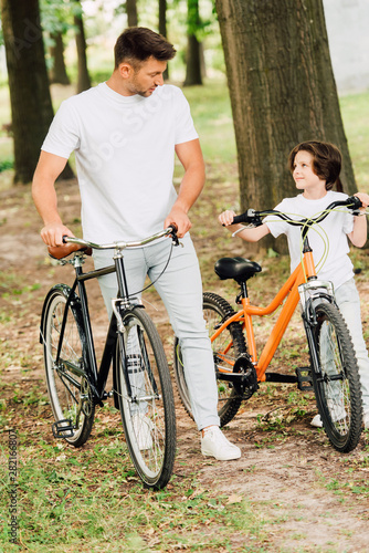 full length view of father and son walking in park with bicycles and looking at each other