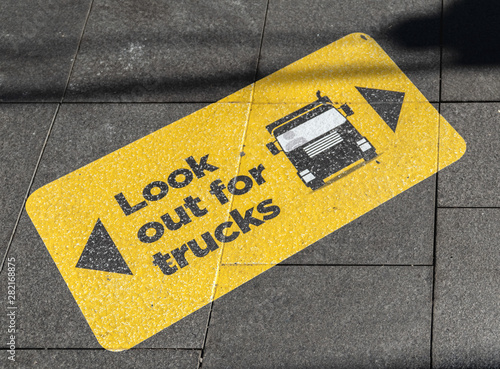 Diagonal view of a yellow and black warning sign stuck on to the pavement near a pedestrian crossing with text, "Look out for trucks" and an image/vector of a truck.