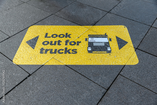 Yellow and black warning sign stuck on to the pavement near a pedestrian crossing with text, "Look out for trucks" and an image/vector of a truck.