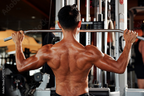 Rear view of strong man working on his back muscles in pull down machine