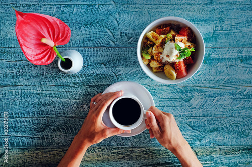 Woman's hands holding cup of coffee. Healthy breakfast with fruit salad bowl and red flower. Top view photo