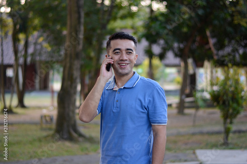 Portrait of handsome Asian male in blue t-shirt talk at phone in outdoor parks © SVRSLYIMAGES